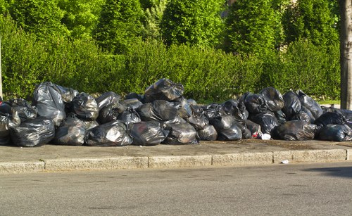 A long row of tightly packed black rubbish bags, made of plastic with a shiny, crinkled texture, are lined up along the edge of a paved street. The bags are placed directly on the sidewalk, with some slight irregularities in their stacking. Behind the row of rubbish bags, there is a dense green backdrop of trees and bushes, indicating a natural environment typical of a suburban or semi-urban area in Whitechapel. The sunlight creates subtle shadows on the bags, highlighting their uneven surface and the plastic's reflective quality. The scene suggests a collection of waste awaiting removal, fitting within the context of a professional rubbish removal service such as Commercial Waste Removal Whitechapel. The overall setting emphasizes urban street waste management, with a focus on waste containment and environmental surroundings in East London.