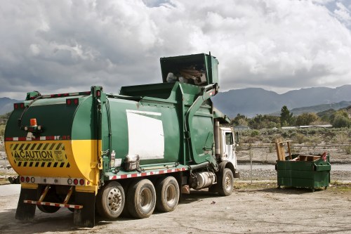 A large green commercial trash collection vehicle parked on an unpaved outdoor area with a cloudy sky overhead. The vehicle features a yellow caution stripe on the rear and a white empty panel on its side, indicating a waste collection body. To the right of the truck, there is a green waste skip filled with various rubbish, situated next to a chain-link fence. In the background, distant mountains and sparse vegetation are visible, suggesting a rural or outskirts setting. The scene is associated with waste removal services, consistent with a professional rubbish collection operation near Whitechapel or a similar London suburb, operated by Commercial Waste Removal Whitechapel, on a day with overcast weather conditions.