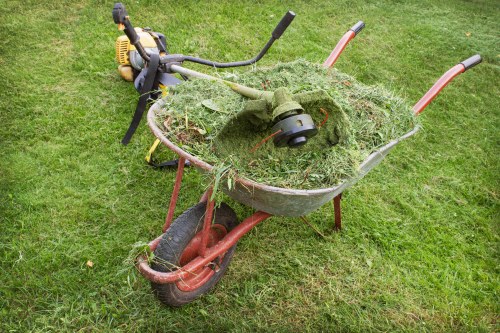 A rusty red wheelbarrow filled with green grass clippings, leaves, and garden debris, positioned on a well-maintained grassy lawn. The wheelbarrow's metal sides are slightly corroded, and it has a black rubber wheel at the front, with two orange handles extending upward at the back. Behind the wheelbarrow, a yellow and black cordless grass trimmer is lying on the grass, with its handle and cutting head partially visible. The background consists of evenly cut, lush green grass, suggesting a residential or community outdoor space. The scene appears to be in daylight, with natural light highlighting the textures of the grass, the metal surfaces, and the gardening tools. The image is relevant to rubbish removal services, such as those offered by Commercial Waste Removal Whitechapel, which specializes in clearing garden and yard waste in the local area around Whitechapel, London.