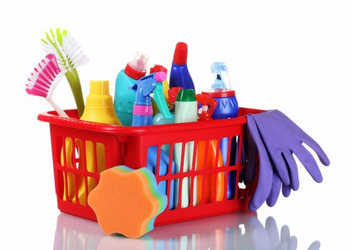 A red plastic basket filled with various cleaning and household supplies is shown, including brightly coloured spray bottles, a green and yellow squeegee, a pink and white cleaning brush, a pair of purple rubber gloves draped over the side, and a cleaning sponge with an orange edge. The items are arranged on a reflective white surface with a plain white background, suggesting a tidy workspace or storage area. The assortment of cleaning products and tools likely relates to waste management or rubbish clearing services offered by Commercial Waste Removal Whitechapel, commonly serving areas around central London, including Whitechapel. The image's focus on cleaning supplies emphasizes domestic or commercial waste clearance tasks, aligning with the company's specialised rubbish removal services. The visual environment appears well-lit, with no clutter or other objects present, fostering a clear view of all items for accessibility and safety considerations related to waste disposal and cleaning operations in the local area.
