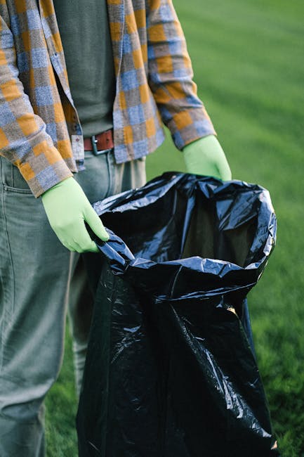 A large pile of black rubbish bags, most of which are tightly tied and bulging with waste, is stacked against a tiled wall in an outdoor urban environment. Some of the bags are torn or partially open,