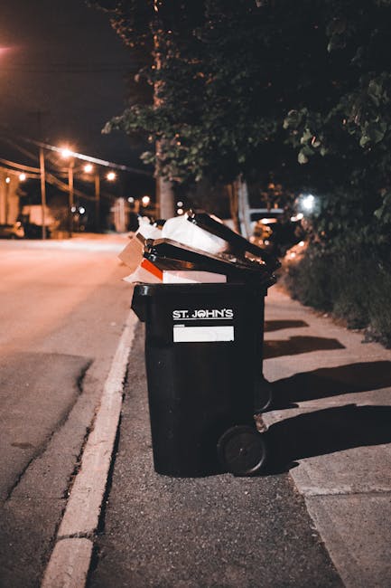 A person is placing a crumpled brown paper bag into the top of a white rubbish bin, which is positioned on a wooden surface indoors. The bin, likely used for waste collection, is partially visible wit